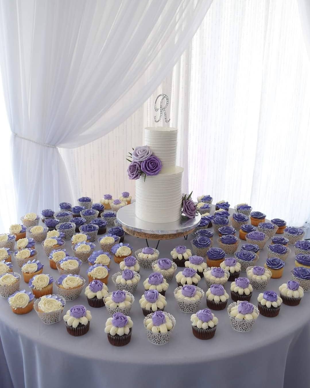White wedding cake with a monogram topper, surrounded by purple and white mini cupcakes